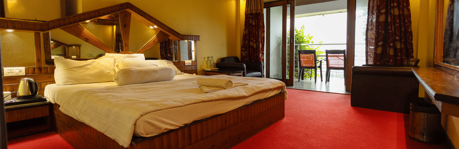 A bedroom with a king size bed, seating area, red carpet, and open balcony door at Hotel Seagull Digha.